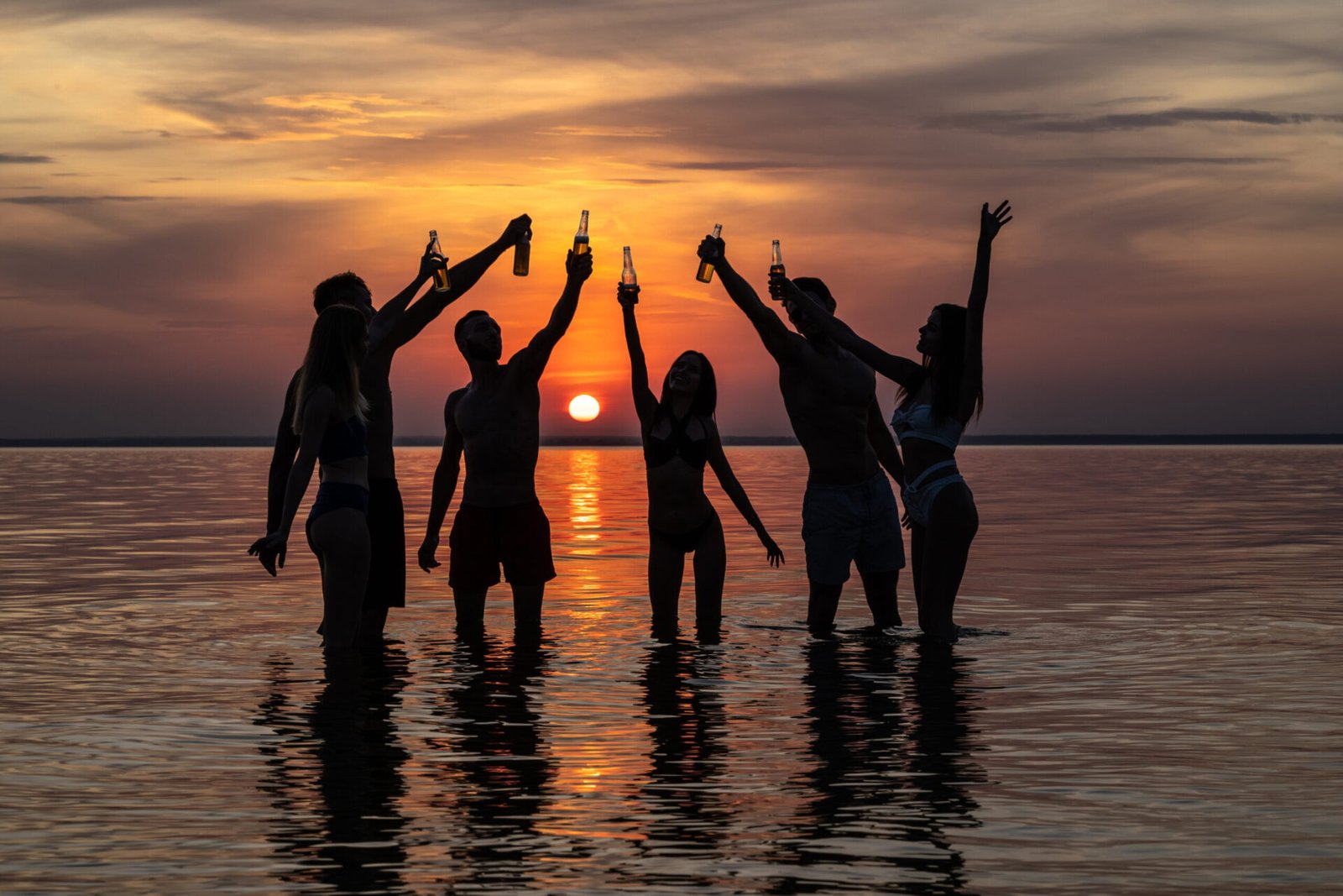 The group of friends standing on the water and making cheers against the sunset