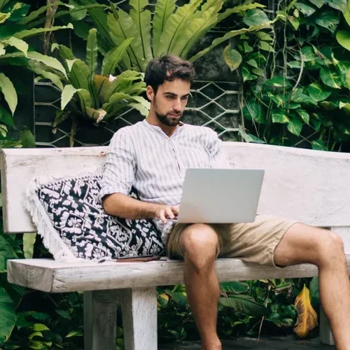 Full body concentrated stylish male in summer clothes surfing internet on netbook while sitting on comfy bench near swimming pool in exoitc resort