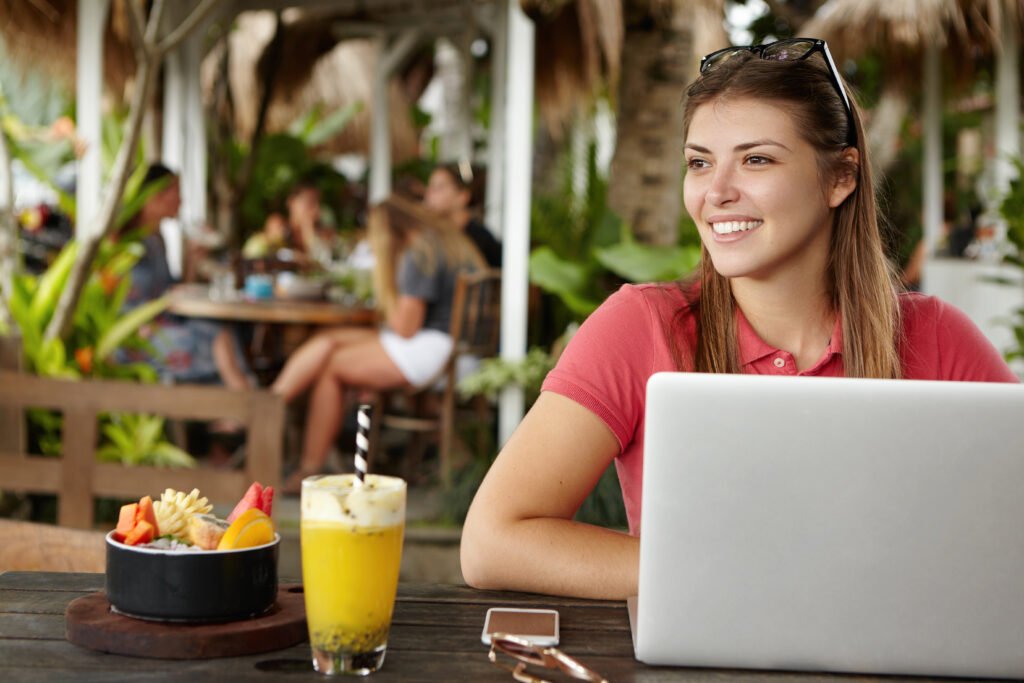 Happy young self-employed woman enjoying free wireless internet connection sitting in front of generic laptop at outdoor cafe. Joyful female using notebook computer during lunch at sidewalk restaurant