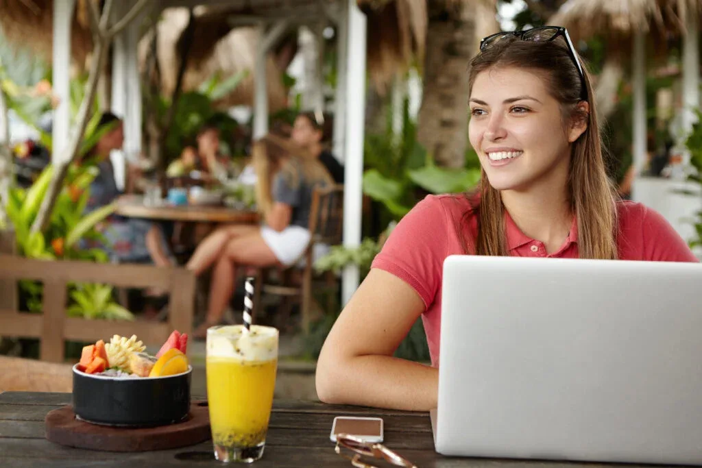 Happy young self-employed woman enjoying free wireless internet connection sitting in front of generic laptop at outdoor cafe. Joyful female using notebook computer during lunch at sidewalk restaurant
