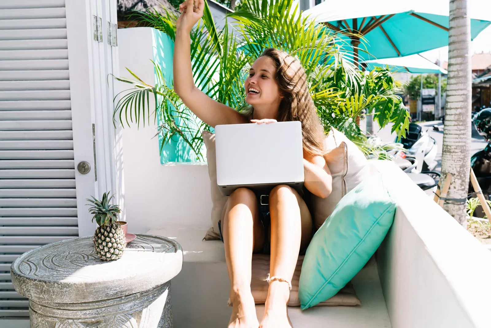 Joyful female in casual clothes smiling and looking away while sitting on terrace with laptop and waving hand on Bali