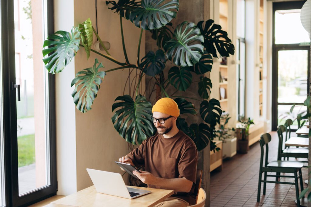 Smiling man wearing yellow hat wipe digital tablet using laptop sitting in modern co working