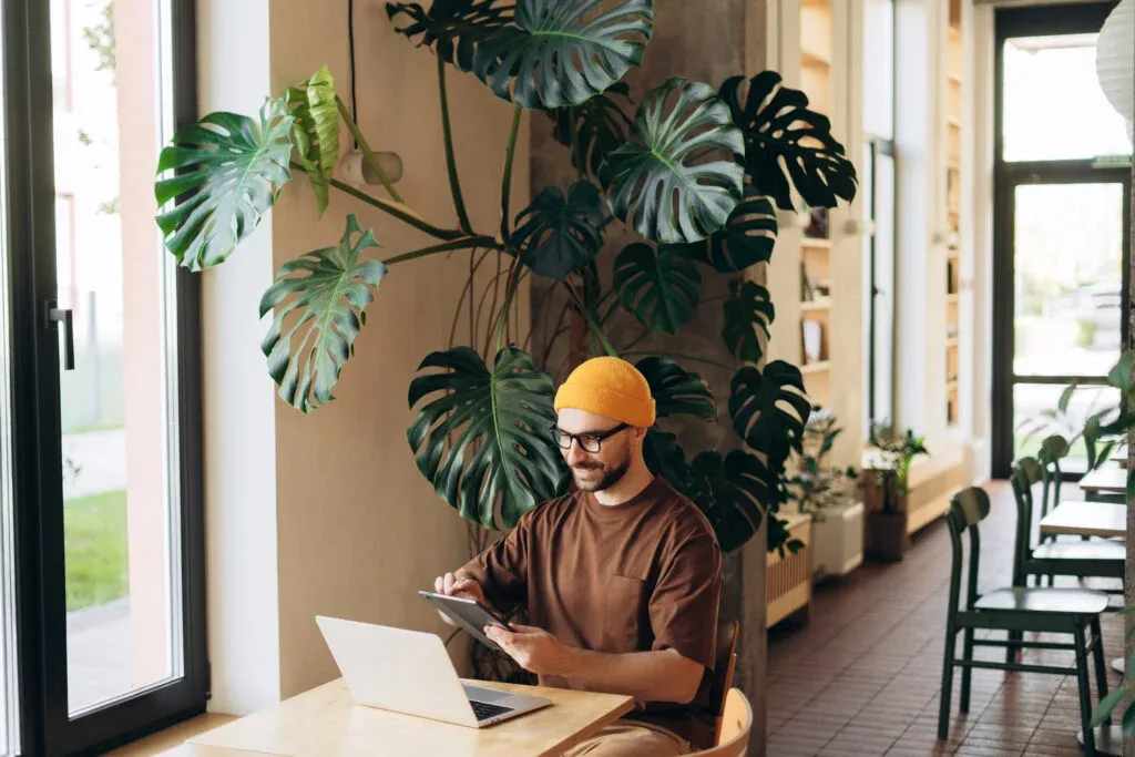 Smiling man wearing yellow hat wipe digital tablet using laptop sitting in modern co working