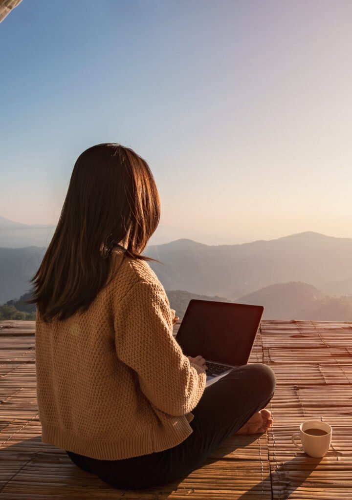 Young woman freelancer traveler working online using laptop and enjoying the beautiful nature landscape with mountain view at sunrise