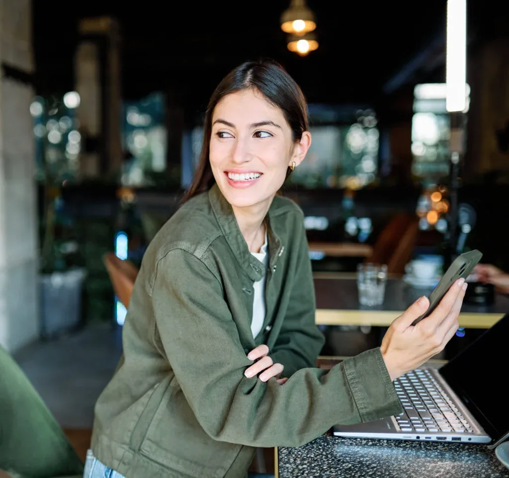 Woman smiling, holding coffee while working on a laptop and using a mobile phone in a cafe